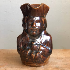 May include: A brown ceramic pitcher shaped like a person wearing a hat. The pitcher has a glazed finish and is detailed with facial features.