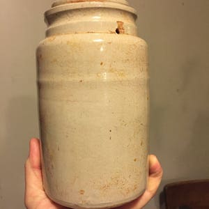 May include: A vintage, off-white stoneware jar with a cylindrical body and a slightly flared rim. The jar has a crackled glaze and a small hole near the top. The jar is held in a person's hand. The jar is approximately 8 inches tall.