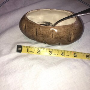 May include: A small, brown ceramic dish with a speckled finish, approximately 4 inches long, with a white interior. A silver spoon rests inside the dish. The dish is on a white surface.