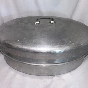 May include: Oval-shaped, aluminum roasting pan with a matching lid. The lid has a curved handle for easy lifting. The pan and lid have a brushed metal finish, showing signs of use. A classic kitchenware item.