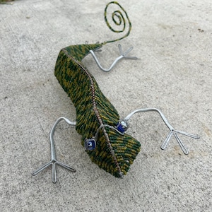 May include: A handcrafted lizard sculpture made of green and yellow beads, with wire legs and a curled tail. The lizard has blue eyes and is lying on a concrete surface. The sculpture is a unique piece of art.