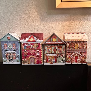 May include: Four decorative house-shaped tins with detailed illustrations. The houses feature various colors, including blue, red, and gingerbread-style brown. Each tin has a unique design, with snow-covered roofs and festive decorations, perfect for holiday storage.