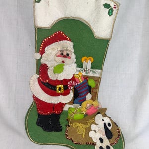 May include: A felt Christmas stocking with a green base and white top. Santa Claus is featured, holding a candy cane. The stocking is decorated with a fireplace, gifts, and a dog. Gold trim and sequins add sparkle to this festive holiday decoration.