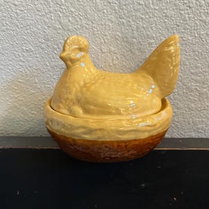 May include: A ceramic hen-shaped container with a lid. The hen is a light yellow color, and the base is a darker brown. The container is likely used for storing eggs or other small items. The design is rustic and charming.
