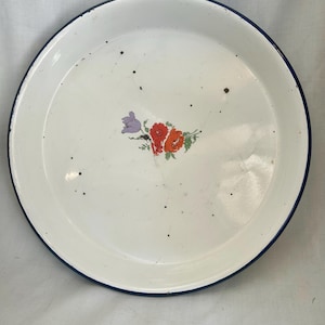 May include: White enamelware tray with a blue rim and floral design. The tray features a cluster of red, orange, and purple flowers in the center. The tray has a circular shape and is likely vintage.