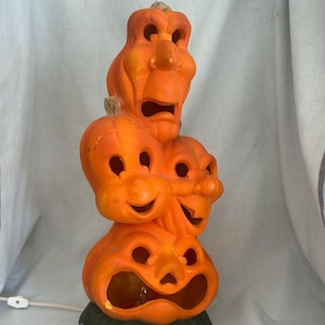 May include: A stacked orange Halloween pumpkin lamp with four carved faces, each with a unique expression. The lamp has a green base and a white power cord. The faces include a surprised, a sad, and two smiling faces.