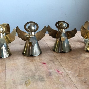 May include: Set of four gold metal angel napkin rings. Each angel has a halo, wings, and a bell-shaped skirt. The angels are holding a small, round object in their hands.