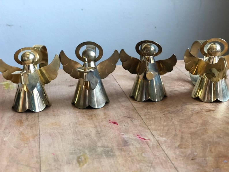 May include: Set of four gold metal angel napkin rings. Each angel has a halo, wings, and a bell-shaped skirt. The angels are holding a small, round object in their hands.
