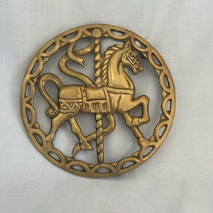 May include: A gold-colored, circular carousel horse wall decor. The horse is in a galloping pose, with intricate details. The horse is surrounded by a decorative, openwork border. The piece is likely made of metal.