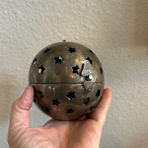May include: A silver-colored, spherical container with star-shaped cutouts. The container is held in a hand and has a hinged lid with a clasp. The surface has a slightly tarnished appearance.