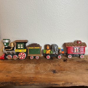 May include: A vintage toy train set with a black locomotive, a green boxcar with "TOOT TOOT" written on it, a flatcar with a lion and elephant, and a red caboose. The train is made of plastic and has a whimsical, cartoonish style.