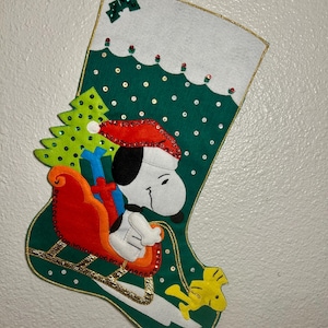 May include: A Christmas stocking featuring Snoopy in a Santa hat, riding a red sleigh with gifts. The stocking is green with white accents, a gold trim, and a small Christmas tree. The stocking is decorated with sequins and a yellow bird.