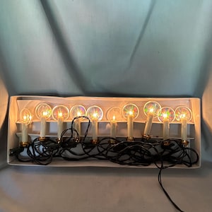 May include: A set of electric candle lights in a white box. The candles are ivory-colored with gold bases and clear, round, iridescent reflectors. Black wires connect the candles. The lights are turned on, emitting a warm glow.