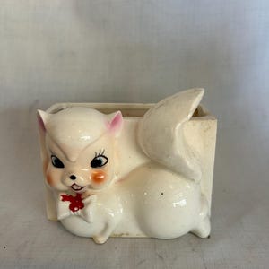 May include: A vintage ceramic planter in the shape of a squirrel. The planter is off-white with pink accents on the ears and cheeks. It features a red bow tie and black eyes. The tail forms part of the planter's structure.