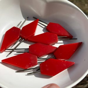 May include: A collection of red, diamond-shaped cocktail forks with silver prongs, resting in a white bowl. The forks have a glossy finish and are arranged in a circular pattern. The bowl is white and has a simple, clean design.