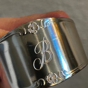 May include: A silver-colored napkin ring with a floral design and the letter "B" engraved on the front. The ring has a curved shape and is made of a shiny metal. The floral design is repeated on the top and bottom of the ring.