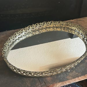 May include: Oval-shaped vanity tray with a mirrored surface and an ornate, gold-colored filigree border. The tray sits on a wooden surface. The decorative tray is suitable for displaying jewelry or perfumes.