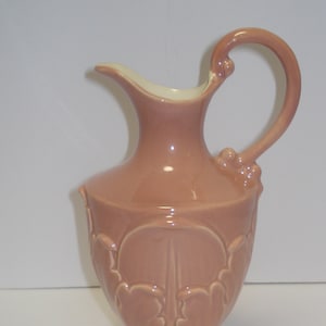 May include: A pink ceramic jug with a decorative floral design and a handle. The jug has a wide base and a narrow neck.