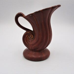 May include: A maroon-colored ceramic pitcher with a unique, swirling design. The pitcher features a handle and a flared spout. The base is a rounded, pedestal shape. The overall design is reminiscent of a seashell.