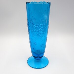 May include: A tall, blue glass vase with a grapevine design. The vase has a flared top and a round base.