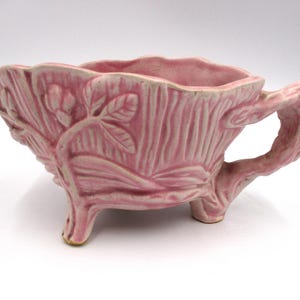 May include: A pink ceramic planter with a tree trunk design and three legs. The planter has a leafy design and a handle.