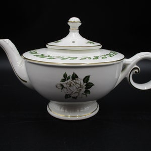 May include: White porcelain teapot with a gold trim and a floral design featuring white roses. The teapot has a lid with a small hole in the top and a decorative handle.