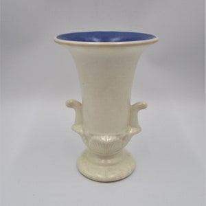 May include: A white ceramic vase with a blue rim and ornate handles. The vase has a classic urn shape with a flared top and a round base.