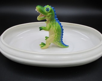 May include: A green and blue plastic dinosaur toy standing on a white oval dish.