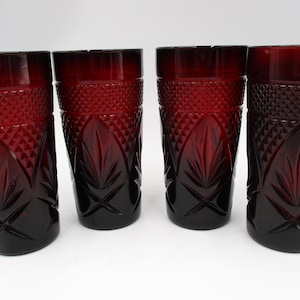 May include: Four ruby red drinking glasses. Each glass has a diamond pattern around the top and a leaf design in the center. The glasses are made of a dark red glass and are empty.