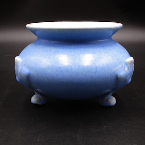 May include: A light blue ceramic pot with three feet and a rounded body. The pot has a smooth, glazed finish.