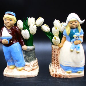 May include: A pair of ceramic Dutch boy and girl figurines, each holding a bouquet of white tulips. The boy is wearing a blue and brown sailor outfit and holding a small sailboat. The girl is wearing a blue and white dress and a white bonnet and is holding a small doll.