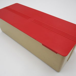May include: A rectangular storage box with a red lid. The lid has a raised design with three parallel lines. The box is made of plastic and has a beige color.