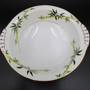 May include: White porcelain serving bowl with a gold rim and handles. The bowl features a bamboo design with green leaves and yellow stalks. The handles have a white and gold pattern.