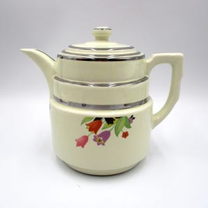 May include: Cream-colored ceramic teapot with a lid and handle. The teapot features silver trim and a floral design with red, purple, and pink flowers. A classic piece for serving tea.