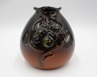 May include: A small, brown ceramic vase with a glossy finish. The vase features a decorative relief of stylized berries and leaves. The top and upper portion of the vase are dark brown, transitioning to a lighter brown at the base. The vase has a rounded shape.
