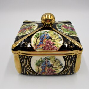 May include: A square ceramic trinket box with a gold lid and a gold knob. The box is decorated with black and gold trim and four panels depicting scenes of women in traditional Japanese clothing.