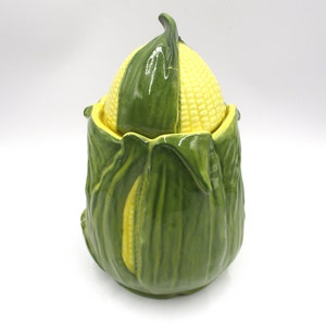 May include: A ceramic corn on the cob cookie jar with a green husk and yellow kernels. The lid is shaped like a corn cob and fits snugly on top of the jar.