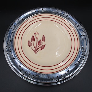 May include: A white ceramic plate with a red floral design, surrounded by a silver-plated rim with an openwork design.