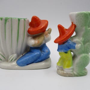 May include: Two vintage ceramic figurines. One is a white vase with green accents and a figure in a red hat and blue pants. The other is a cactus-shaped vase with a similar figure. Both are painted in bright colors.