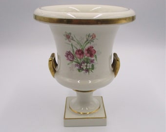 May include: A white ceramic urn with gold accents and a floral design. The urn has two gold handles and sits on a square base.