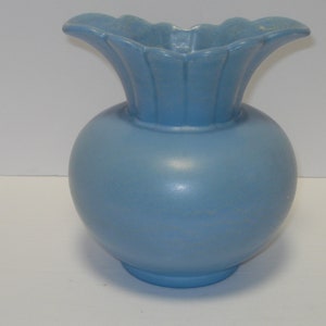 May include: A light blue ceramic vase with a wide, ruffled opening. The vase has a round body and a smooth, matte finish.