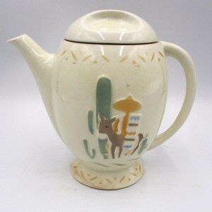 May include: Cream ceramic teapot with lid, spout, and handle. The teapot has a desert theme with a cactus, donkey, and other southwestern elements. The base and rim have a decorative pattern.
