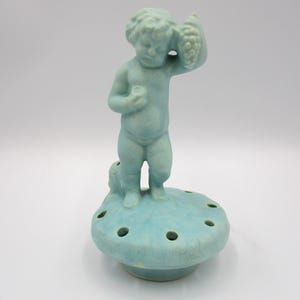 May include: A light blue ceramic figurine of a cherub holding grapes, standing on a round base with several holes. The figurine is a vintage piece, likely used as a flower frog. The base is approximately 4 inches in diameter.