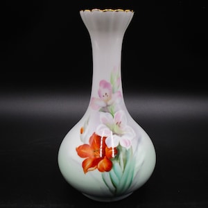 May include: A white porcelain vase with a gold rim and a floral design. The vase features pink and orange flowers with green leaves.