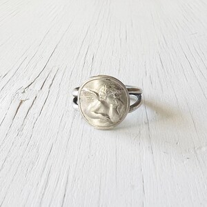 May include: Silver ring with a round, raised design of an angel with wings. The angel is holding its face in its hands.