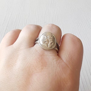 May include: A silver ring with a round design featuring a cherub with wings. The ring is worn on a person's finger.