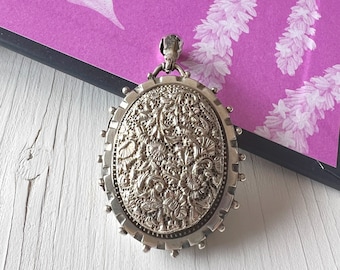 May include: Silver oval pendant with detailed floral and leaf patterns. The pendant features a decorative edge and a small loop for a chain. It is shown on a white wooden surface with a purple background.