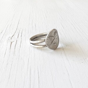 May include: A silver ring with a double band and a round, textured design featuring an angel figure.