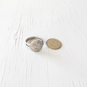 May include: Silver ring with a double band and a round design featuring a winged angel. The ring is shown next to a Canadian one dollar coin for size comparison.