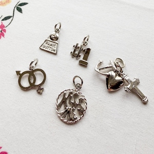 May include: A selection of silver charms. The charms include a "Panic Button", a number one, a heart with an anchor and cross, a gender symbol, and a charm with the word "Mrs" inside a circle.